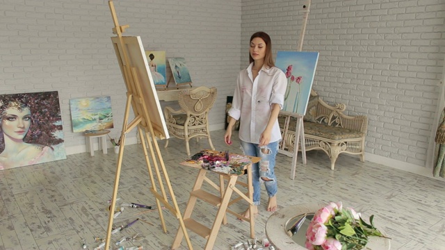 一个穿着白衬衫的年轻性感的女孩在画室的画布上画画。艺术视频素材