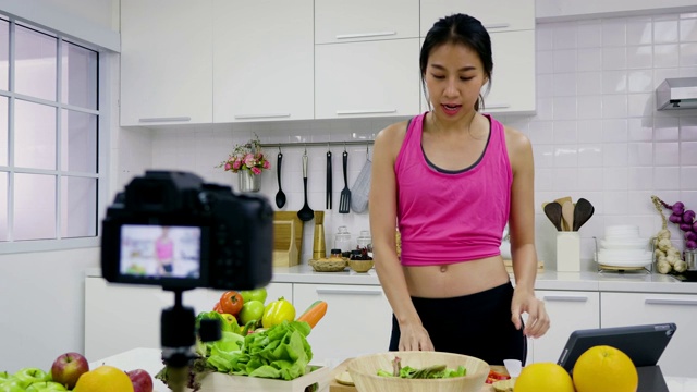 年轻的亚洲运动女性美食博主聊天，同时录制视频准备绿色沙拉在厨房，教程视频到社交网络通过互联网在家概念视频素材