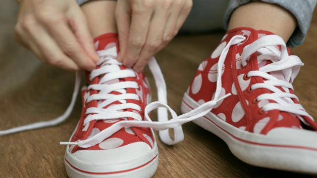 年轻女子坐在地板上解开红色球鞋的鞋带。特写女孩的手系鞋带准备运动，走在街上。视频素材