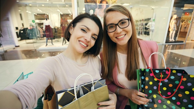 年轻的女学生们在购物中心自拍，展示了明亮的纸袋和竖起的大拇指，做着滑稽的鬼脸。购物狂和joy概念。视频下载