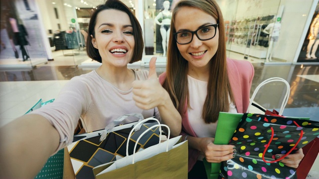 快乐的朋友从购物中心在线视频通话的观点。女孩们拿着电子设备，看着相机聊天，展示纸袋和大拇指。视频下载