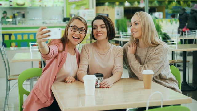 年轻漂亮的女士们坐在咖啡馆的桌子旁用智能手机自拍，摆姿势喝饮料。友谊、休闲时间和现代科技理念。视频素材