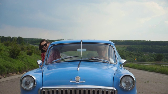 一对年轻夫妇开着老爷车在乡村公路上旅行。女孩探出窗外的复古汽车和享受旅行。美丽的风景作为背景。暑假旅行。前视图慢动作视频素材
