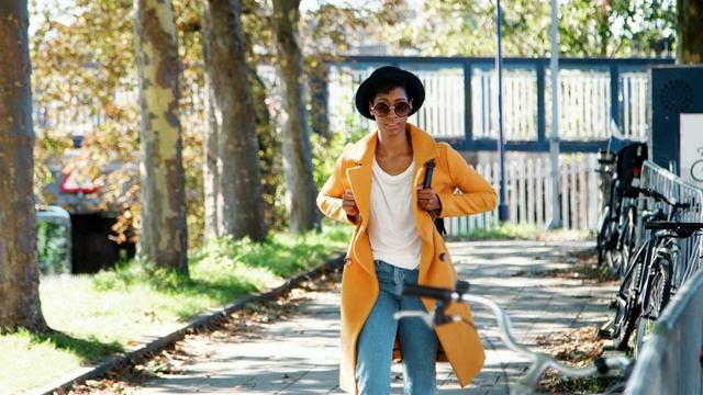 时尚的年轻黑人妇女戴着帽子，太阳镜，蓝色牛仔裤和黄色的豌豆大衣走在一个阳光明媚的日子里，看着周围，微笑着，正面的观点视频下载
