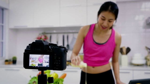年轻的亚洲运动女性食品博主说话，同时录制视频准备绿色沙拉在厨房，教程视频到社交网络通过互联网在家概念，选择性焦点视频素材