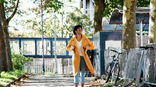 时尚的年轻黑人妇女戴着一顶帽子，太阳镜，蓝色牛仔裤和一件黄色的豆皮大衣走在一条有树的街道上，对着摄像头，用耳机听音乐，环顾四周，背光，低角度视频素材