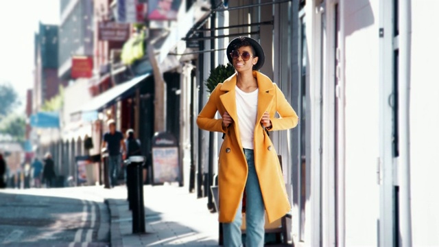 阳光明媚的日子里，穿着蓝色牛仔裤和一件敞开的黄色豆蔻大衣的时尚年轻黑人妇女微笑着走在商店附近的人行道上视频下载