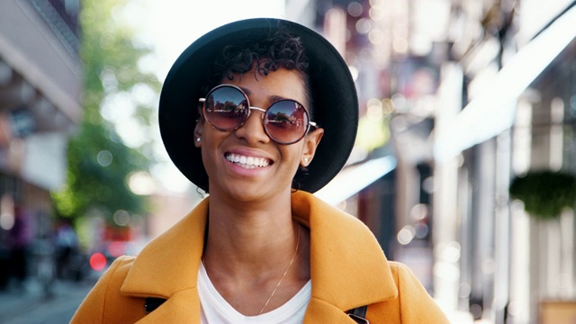 千禧一代的黑人妇女穿着黄色的豌豆外套，太阳镜和洪堡帽，站在城市的街道上对着镜头微笑，特写视频下载