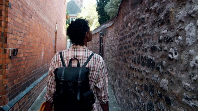千禧一代黑人妇女穿着格子衬衫背着背包走在一个历史街区的狭窄巷子里，环顾四周，后视镜，跟随射击视频下载