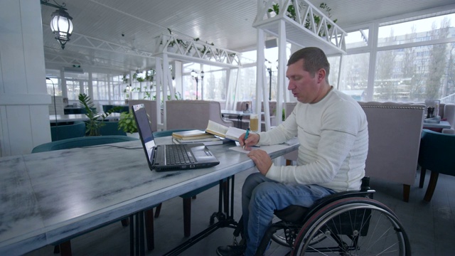 在线教育，坐在轮椅上的残疾学生利用现代笔记本电脑技术从在线课程和书中学习，在咖啡馆的桌子上近距离地在笔记本上做笔记视频下载