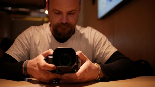 一个留着胡子的男人正坐在咖啡馆的桌子旁，手里拿着照相机视频下载