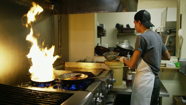 十几岁的西班牙裔男性厨师在一家墨西哥餐厅准备三个平底锅肉和蔬菜在一个商业范围内与火焰视频下载