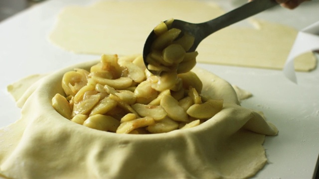 一个人在做苹果派的时候，用一个大的金属勺子把煮熟去皮的苹果片(馅料)从有盖的烤盘里舀到包着糕点的馅饼罐里视频下载