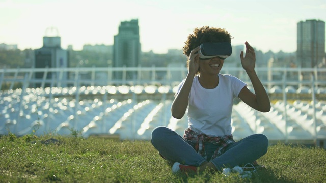 有趣的卷发女性使用VR头盔，坐在草坪上，在阳光明媚的日子，小玩意视频素材