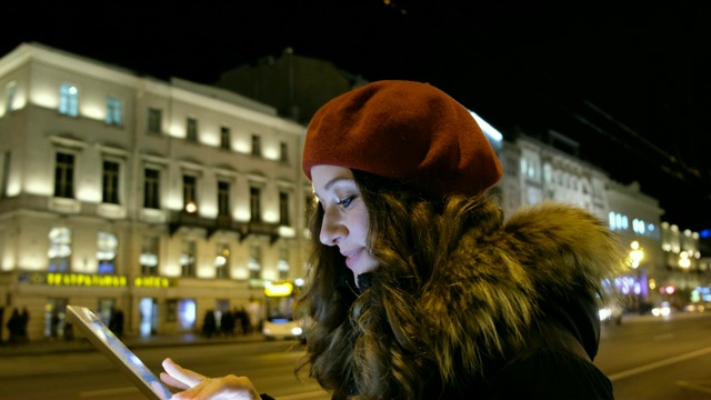 年轻女孩用平板电脑看着屏幕和手指和微笑。在夜间城市的背景下，移动的汽车和行走的人群视频素材