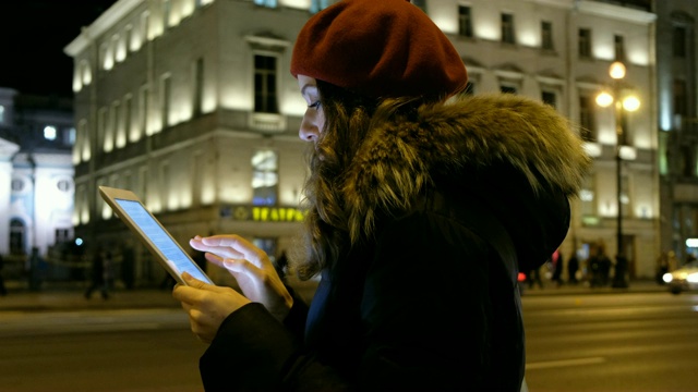 年轻女孩用平板电脑看着屏幕和手指和微笑。在夜间城市的背景下，移动的汽车和行走的人群视频素材