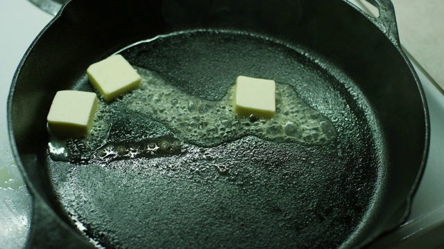 一个女人的手把一小块黄油丢进炉子上的黑色铸铁长柄锅里，然后在黄油融化时用木铲搅拌它们视频下载