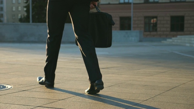 一个年轻商人的脚拿着公文包走在城市的街道上。通勤上班的商人。穿着西装的自信男人正在上班的路上。城市景观背景。慢镜头后视图特写视频下载