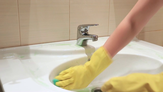 一个戴着黄色橡胶手套的不可辨认的女人正在用绿色海绵清洗浴室的水槽。清洁和洗涤概念视频素材