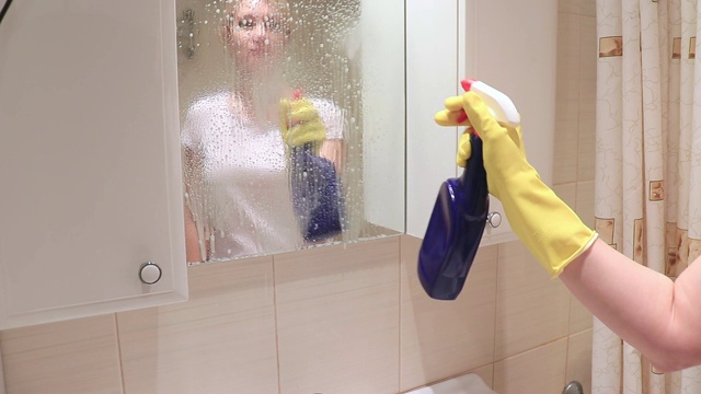 年轻的金发女人戴着黄色橡胶手套在擦洗浴室的镜子，洒上雨刷喷雾和泡沫清洁剂。清洁、擦拭和清洗镜子的概念视频素材