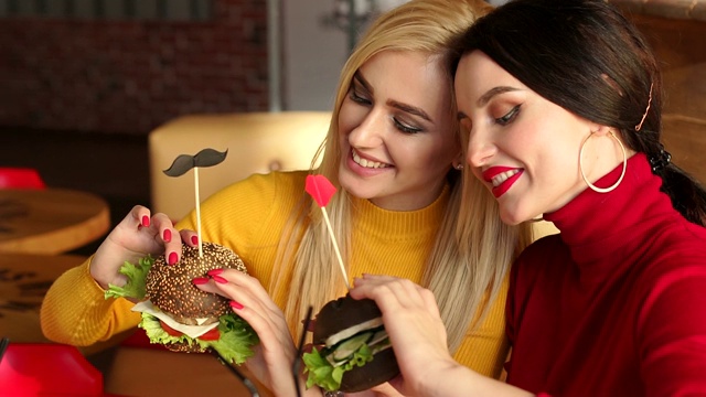 两个漂亮的年轻女孩和明亮的化妆在咖啡馆吃汉堡。慢动作视频素材