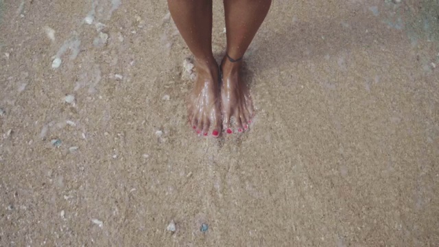 特写慢动作拍摄的女性的脚站在沙滩上，海浪溅起，洗走了沙子视频下载