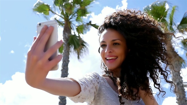 美丽的年轻女子在蓝天和手掌的映衬下用智能手机自拍的慢镜头视频素材