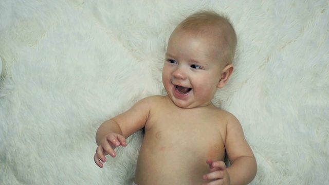 5个月大的男孩躺在一张白色的毯子上，慢镜头中他对着镜头甜甜地笑着。视频素材