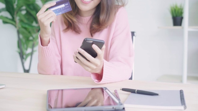 美丽的亚洲女人使用智能手机在网上购物通过信用卡，而穿着毛衣坐在家里客厅的桌子上。生活方式女性在家的概念。视频素材