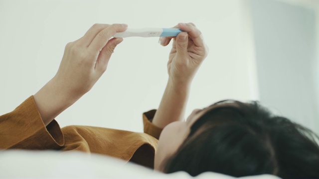 亚洲妇女躺在床上，并持有阳性怀孕测试试剂盒视频素材
