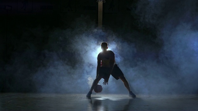 一名男子在黑暗的篮球场上拿着篮球，背景是烟雾中的篮球圈，在三盏背光灯的照耀下，他展示着运球技巧视频下载