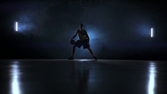 在一个黑暗的房间里，一个篮球运动员在球场上运球，在烟雾中有一个慢动作的背光视频素材