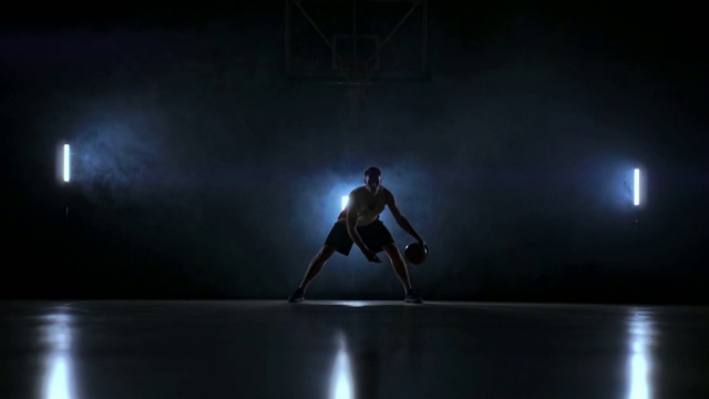 一名男子在黑暗的篮球场上拿着篮球，背景是烟雾中的篮球圈，在三盏背光灯的照耀下，他展示着运球技巧视频素材