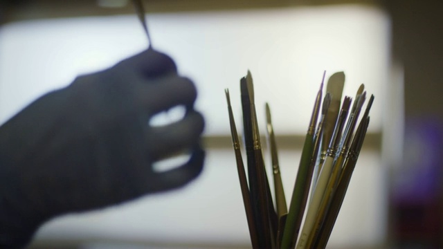 在室内艺术工作室里，一位戴着防护手套的艺术家伸出手从一堆画笔中选择一支画笔视频下载
