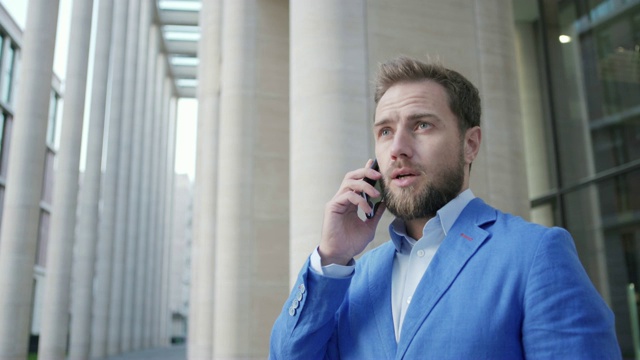 在办公楼入口附近，一个穿着蓝色夹克，满脸胡须，自信的商人正在打手机，中景视频素材