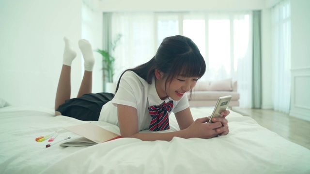 一名穿制服的女学生躺在床上使用智能手机视频下载