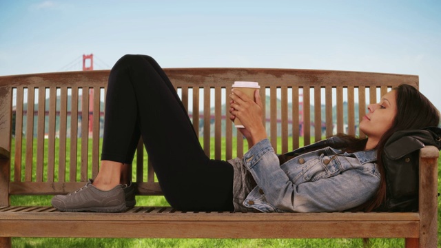 穿着牛仔夹克的时髦女孩躺在金门大桥附近的公园长椅上发短信视频下载