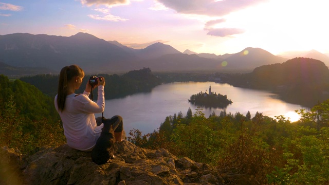 旅行者女孩坐在她的小狗旁边，拍着日落的照片。视频下载
