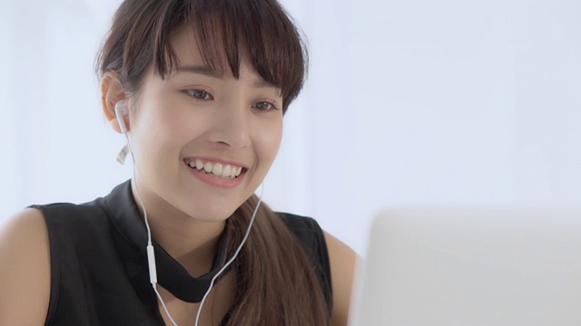 美丽的年轻亚洲女人微笑着坐在用笔记本电脑听音乐与耳机放松在办公室，女孩我们笔记本听广播上网，生活理念。视频素材