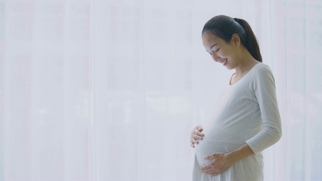 美丽的亚洲孕妇透过窗户看，并在客厅揉胃。视频素材