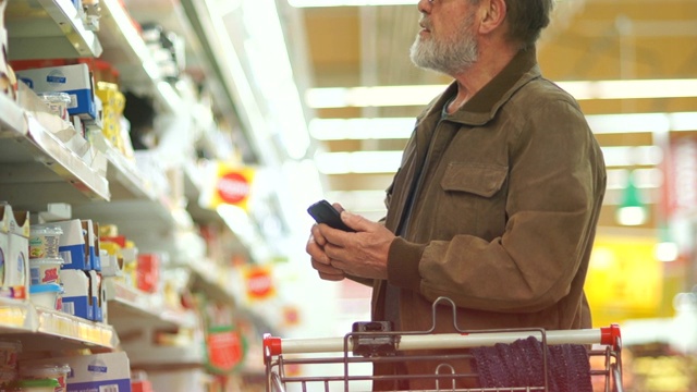 一位留着灰白胡子、戴着眼镜的退休老人在超市里挑了一块奶酪，然后把它放进手推车里。乳制品、健康食品视频素材