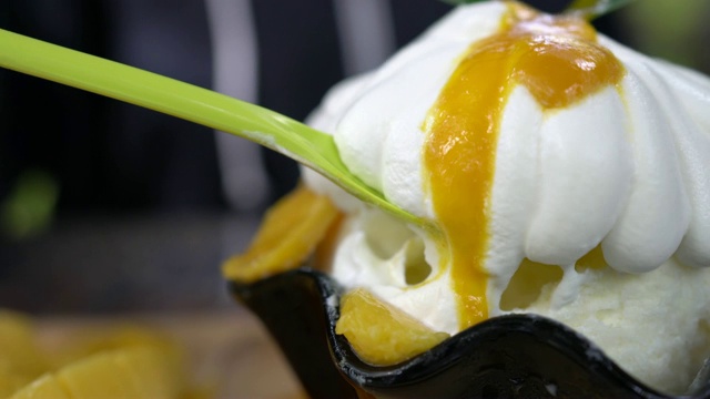 近距离拍摄的长绿色勺子舀白色甜奶油在芒果冰苏。韩国的一种冰甜点，由牛奶刨冰加上甜奶油加上芒果和芒果酱制成。视频素材