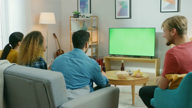 四个朋友坐在家里的沙发上，一边看绿屏电视，一边吃零食和喝饮料。年轻人在家里玩。视频素材