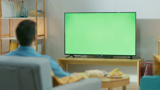 快乐的家伙坐在家里客厅的椅子上，看着绿色色卡屏幕，放松工作后。男人在舒适的房间里看体育比赛，新闻，电视节目或电影。视频素材