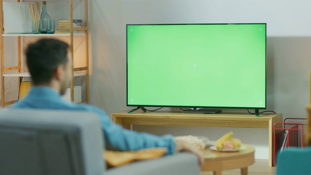 快乐的家伙坐在家里客厅的椅子上，看着绿色色卡屏幕，放松工作后。在舒适的房间里看体育比赛、新闻、电视节目或电影的男人。视频素材