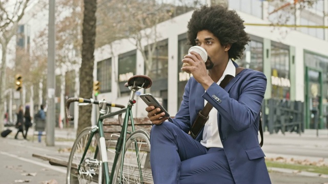 一个商人坐在长凳上，一边喝着咖啡，一边查看手机短信视频素材