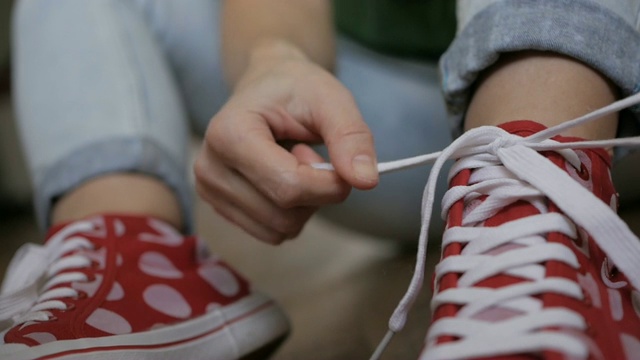 中景镜头。一个女人坐在地板上系着红色运动鞋的鞋带。特写女孩的手系鞋带准备运动，走在街上。缓慢的运动。视频下载