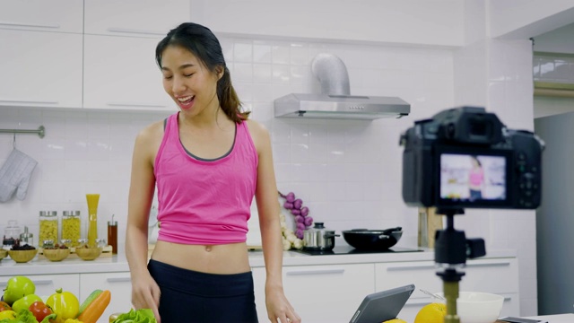 年轻的亚洲运动女性食品博主说话，同时录制视频准备绿色沙拉在厨房，教程视频到社交网络通过互联网在家概念，选择性焦点视频素材