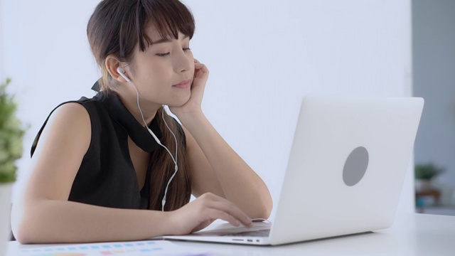 美丽的年轻亚洲女人微笑着坐在用笔记本电脑听音乐与耳机放松在办公室，女孩我们笔记本听广播上网，生活理念。视频素材