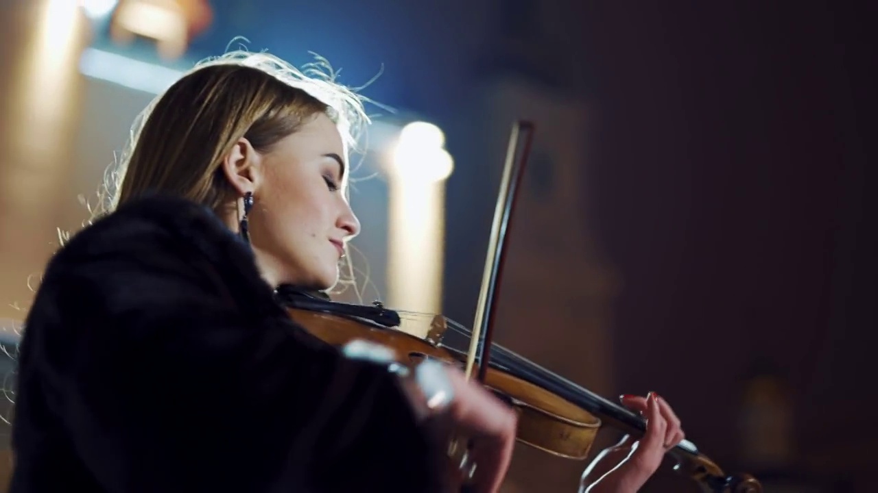 一个穿着黑色外套的富有创造力的女孩正在拉着小提琴，在城市夜晚灯笼的灯光下弹奏着浪漫的旋律。视频素材